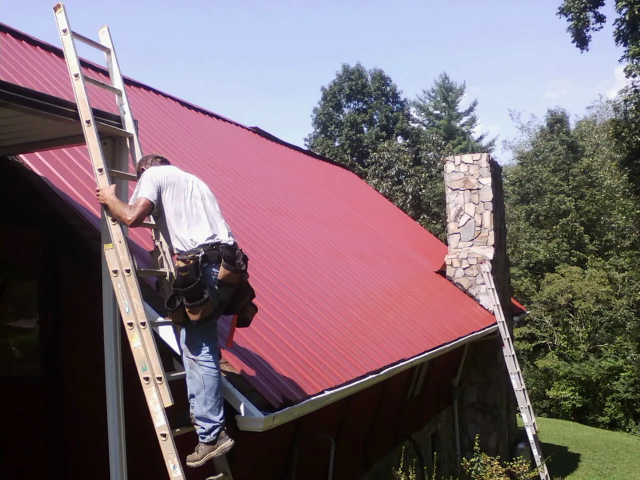 Roofer on a ladder beside a red metal roof and stone chimney — Soffit & Fascia Repair by Roofing Brothers in Ferndale